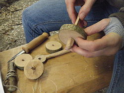 Das Bild zeigt Hände, die einen angespitzten Holzstab durch ein Loch in der Mitte einer Holzscheibe stecken. Auf der Bank auf der die Person sitzt liegen weitere Holzscheiben, ein Messer und ein Holzstück, das mit einer Schnur umwickelt ist. 