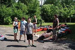 Das Bild zeigt eine Gruppe von wartenden Jungs auf einem asphaltierten Platz an einer Wiese. Auf der Wiese liegen Kanus und andere Utensilien.