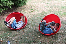 Das Bild zeigt zwei Jungen, die mit angezogen Beinen in zwei roten Plastikschalen, den sogenannten Kuller-Kreiseln, liegen und das Victory-Zeichen machen.