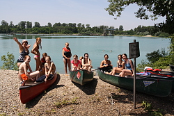 Das Bild zeigt eine Gruppe von Mädchen, die fünf Kanus ans Kiesufer eines Baggersees gezogen hat und in diesen sitzt beziehungsweise neben diesen steht. Das Mädchen, das ganz links steht, reckt jubelnd die Arme in die Luft. 