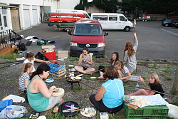 Das Bild zeigt eine Gruppe von Mädchen, die auf einem Hof um einen Grill sitzen und Stockbrot backen. Im Hintergrund sind mehrere Bullis mit einem Kanu-Anhänger zu sehen. Überall verstreut liegen andere Utensilien.
