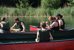 Das Bild zeigt mehrere Kanus auf dem Wasser nebeneinander. Die Insassen tragen Schwimmwesten und unterhalten sich