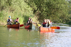 Das Bild zeigt Mädchen, die zu zweit oder zu dritt in drei Kanus auf einem Fluss paddeln. Das vorderste Mädchen zeigt das Victory-Zeichen.