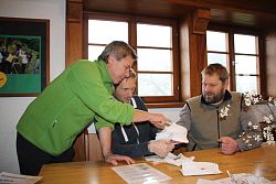 Das Bild zeigt einen Referent und zwei Teilnehmer der Fortbildung "Outdoor-Erste-Hilfe", die in einem Raum an einem Tisch sitzen beziehungsweise stehen. Einem wird am rechten Arm und rechter Hand ein Verband anlgelegt.