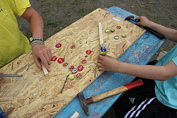 Das Bild zeigt Kinderhände, die Kronkorken auf Holzlatten nageln und so Rasseln herstellen.