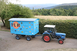 Das Bild zeigt das Spielmobil des Kinder- und Büros, einen hellblau gestrichenen und mit zahlreichen Bildern beklebten Bauwagen, der an einem blauen Traktor hängt.