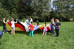 Das Bild zeigt Kinder und Jugendliche, die im Kreis stehen und ein regenbogenfarbiges Fallschirm-Tuch schwingen.