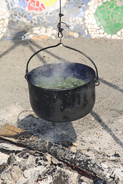 Das Bild zeigt einen Feuer-Topf über einer Feuerstelle. In dem Topf dampft eine Gemüsesuppe.