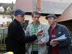 Das Bild zeigt drei männliche Teilnehmer bei der Fortbildung Geocaching des Kinder- und Jugendbüros, die auf ein GPS-Gerät schauen, das der Mittlere um den Hals trägt und der Linke gerade anhebt. Der Rechte hält zudem einen Stapel Papier in der Hand.